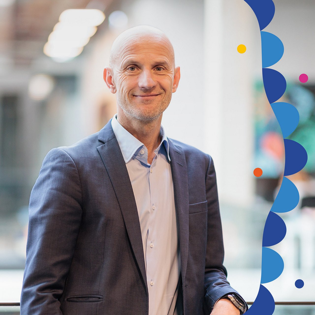 Portrait of John Wittesaele. He is standing in a modern office building, wearing a light blue shirt and a dark blue blazer. He is leaning slightly against a railing with one arm and looking at the camera with a subtle smile. The background is blurred, showing bright structures and colorful wall art.