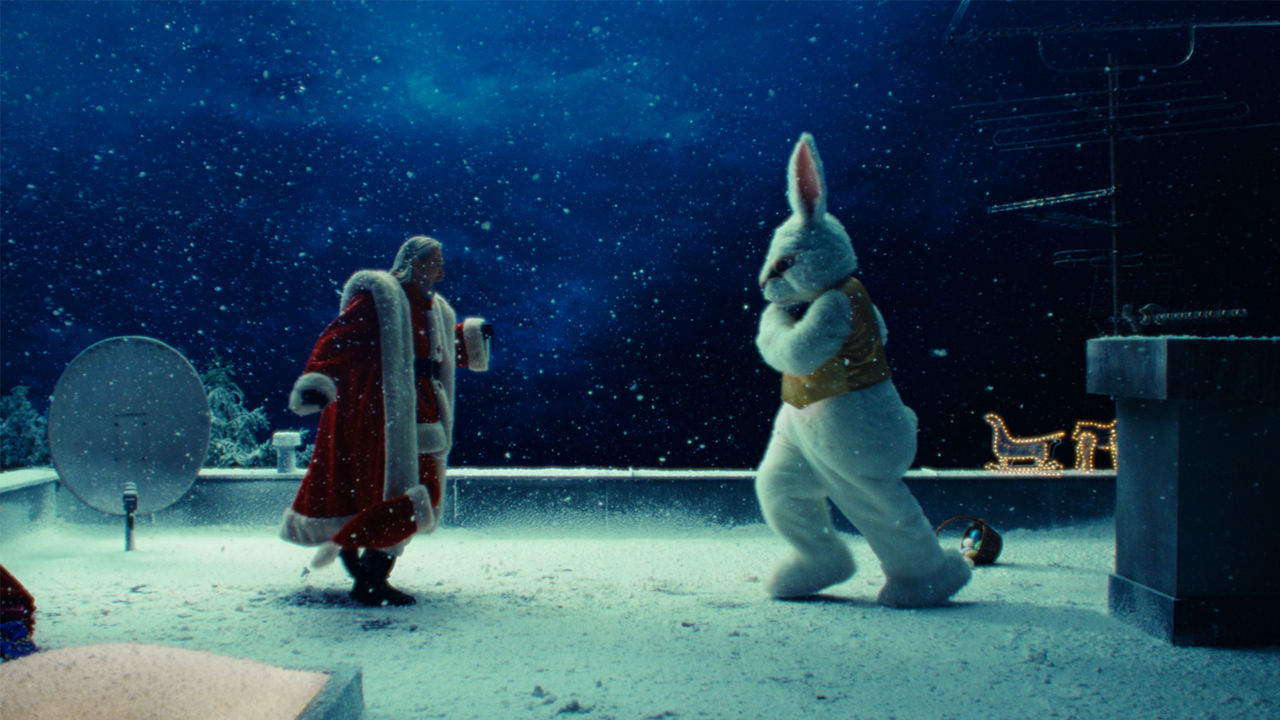 Auf einem verschneiten Dach rennen Weihnachtsmann und Osterhase im Schneetreiben aufeinander zu, als wollten sie kämpfen.