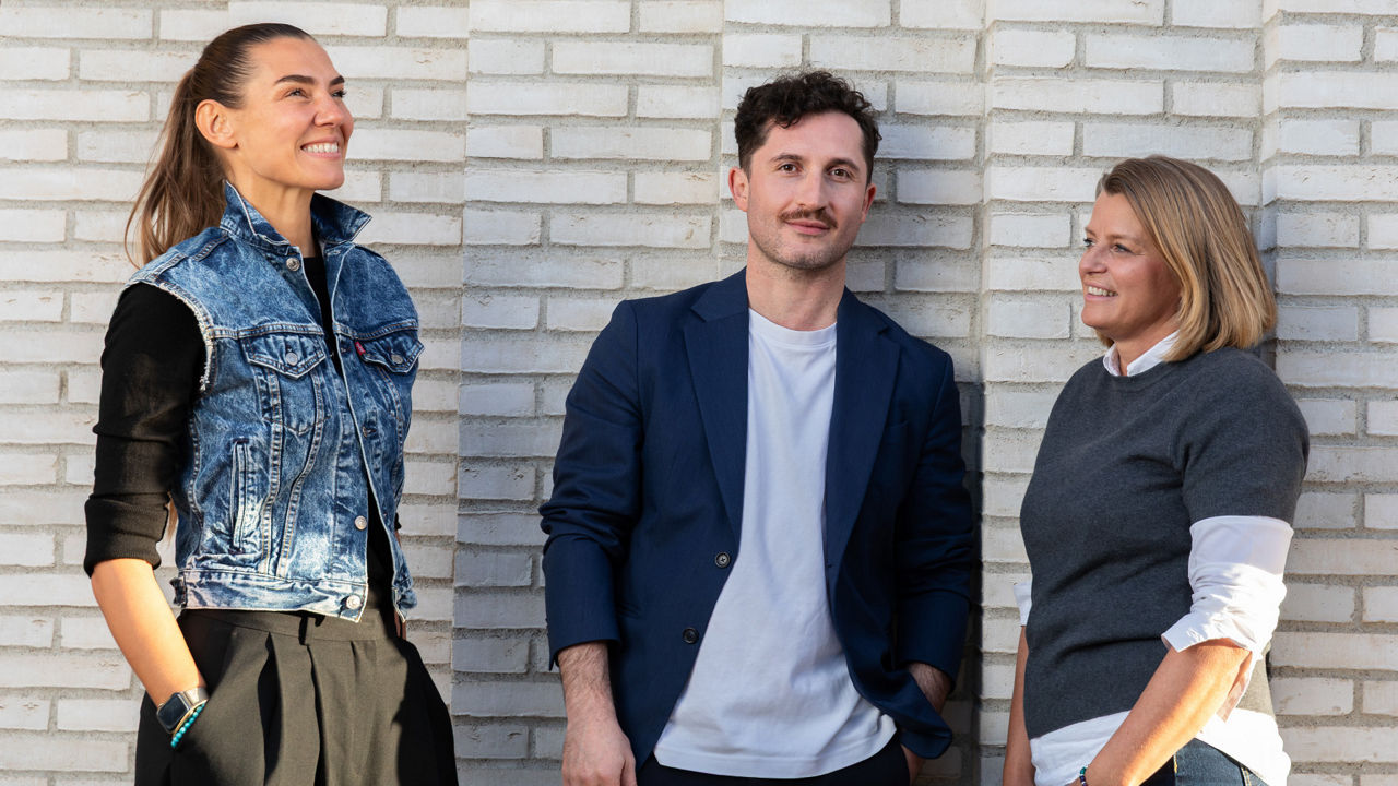 Menschen stehen vor einer hellen Backsteinwand und blicken lächelnd in verschiedene Richtungen. Sie wirken entspannt und freundlich – eine moderne Business-Aufnahme mit natürlichem Licht.