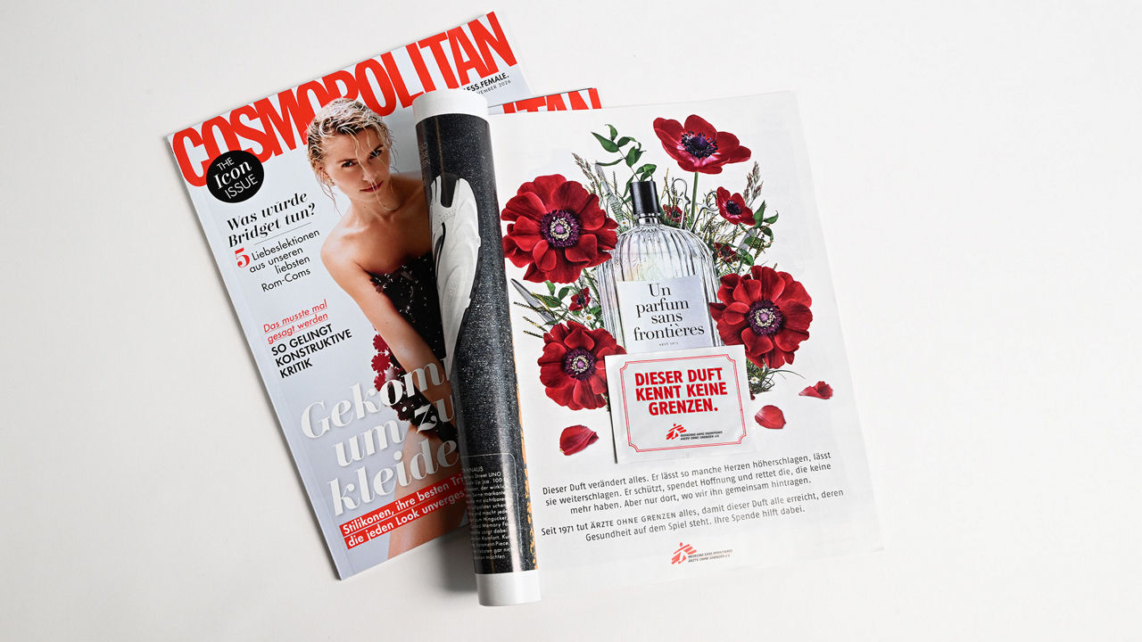 Auf einem hellen Untergrund liegt ein aufgeschlagenes Exemplar des Magazins „Cosmopolitan“. Die rechte Seite zeigt eine Anzeige mit einem Parfümflakon, roten Blumen und dem Schriftzug „Dieser Duft kennt keine Grenzen.“ Im unteren Bereich befinden sich weitere Informationen zur Organisation Ärzte ohne Grenzen. Die linke Seite zeigt das Titelblatt des Magazins mit einer Person auf dem Cover.