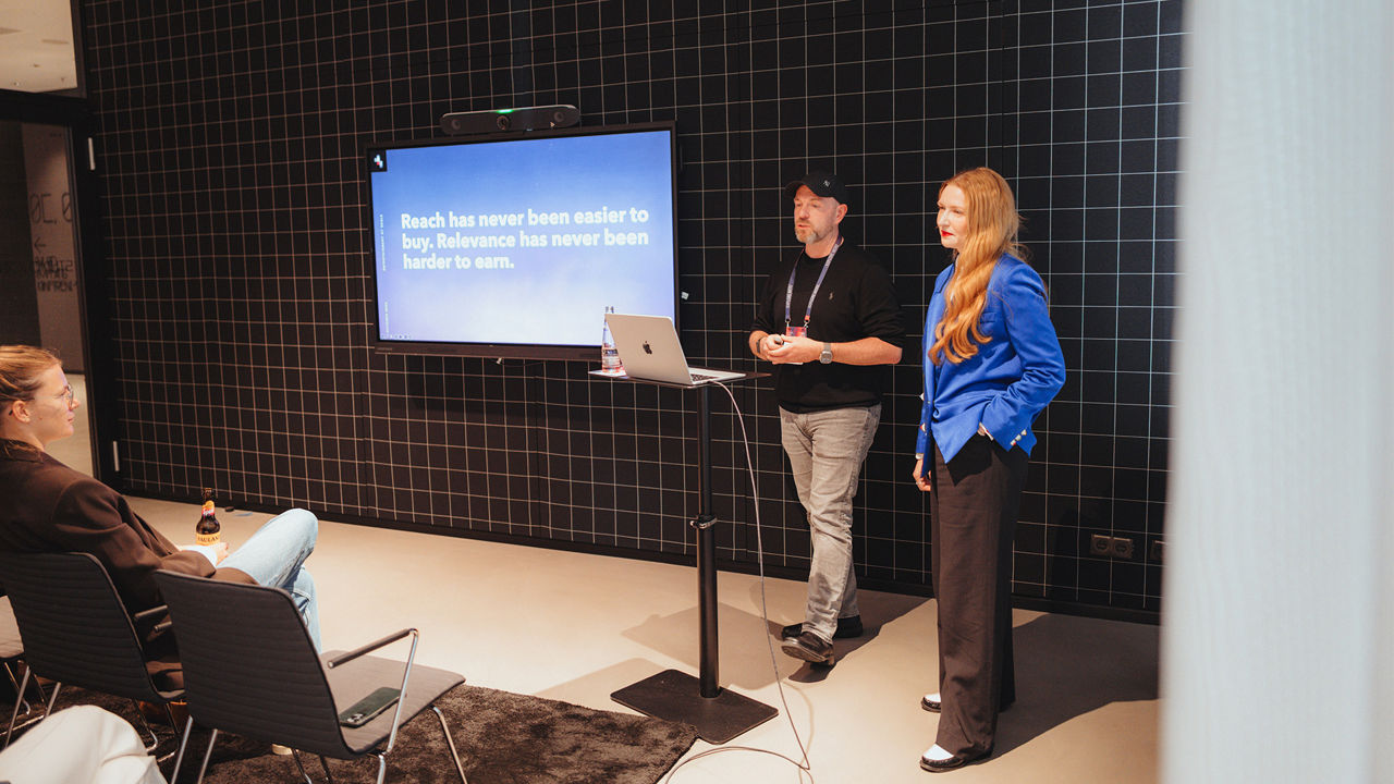 Two people (Alex Turtschan and Franziska Gregor) give a presentation to a seated audience. A standing table with an open laptop is positioned beside the presenters. A large screen in the background reads, “Reach has never been easier to buy. Relevance has never been harder to earn.” The room features a dark, grid-patterned wall.
