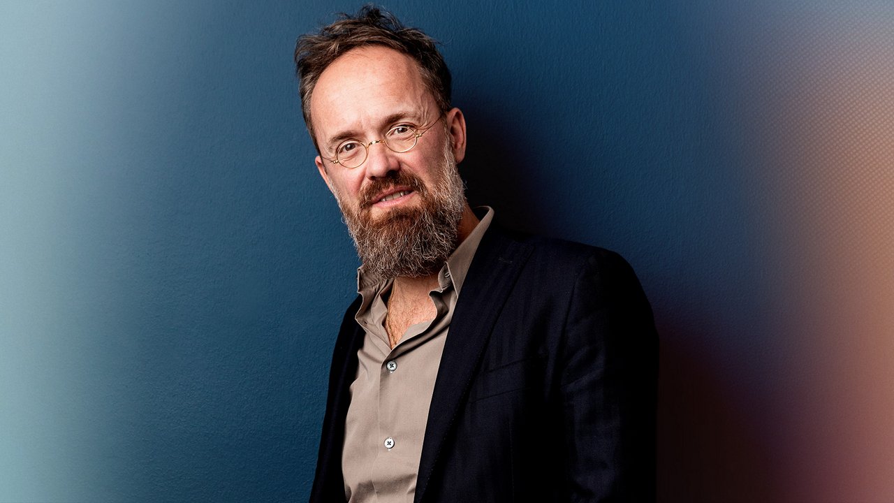 Portrait of Alex Schill against a blue background. He is wearing round glasses, a dark blazer and an open-collar shirt.