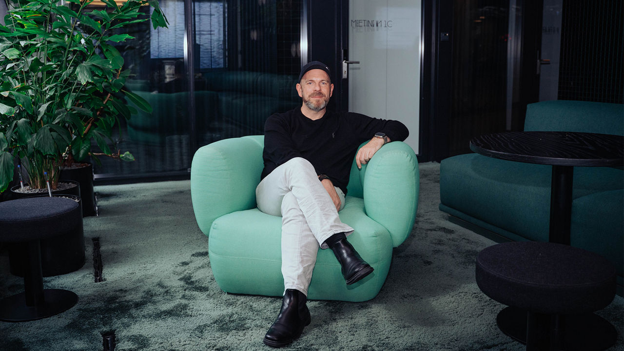 A person (Alex Turtschan) wearing a dark sweater, light-colored pants, and a cap is seated in a light green armchair in a modern office lounge. The room features dark glass walls, green carpeting, indoor plants, and seating areas with tables. The person is facing the camera while seated with crossed legs. The image is presented as a professional environmental portrait in a contemporary workplace setting.