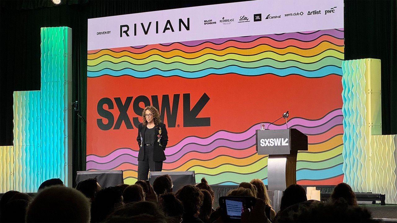 A speaker stands on stage at the SXSW conference in front of a large colorful SXSW backdrop while the audience watches.