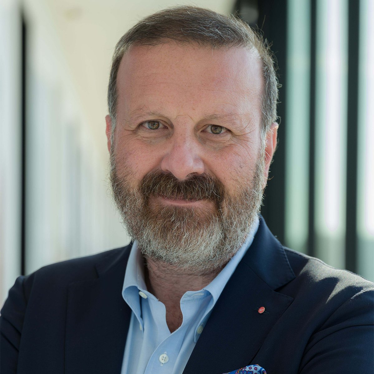 Andrea Michelangelo Pacella: Ein Porträtfoto eines Mannes mittleren Alters mit kurzem Haar und Vollbart, der lächelt und die Arme verschränkt. Er trägt ein dunkelblaues Sakko mit einem bunten Einstecktuch, ein hellblaues Hemd und eine Armbanduhr mit silbernem Metallarmband. Der Hintergrund zeigt einen hellen, modernen Büroflur mit großen Fenstern, durch die Tageslicht einfällt.