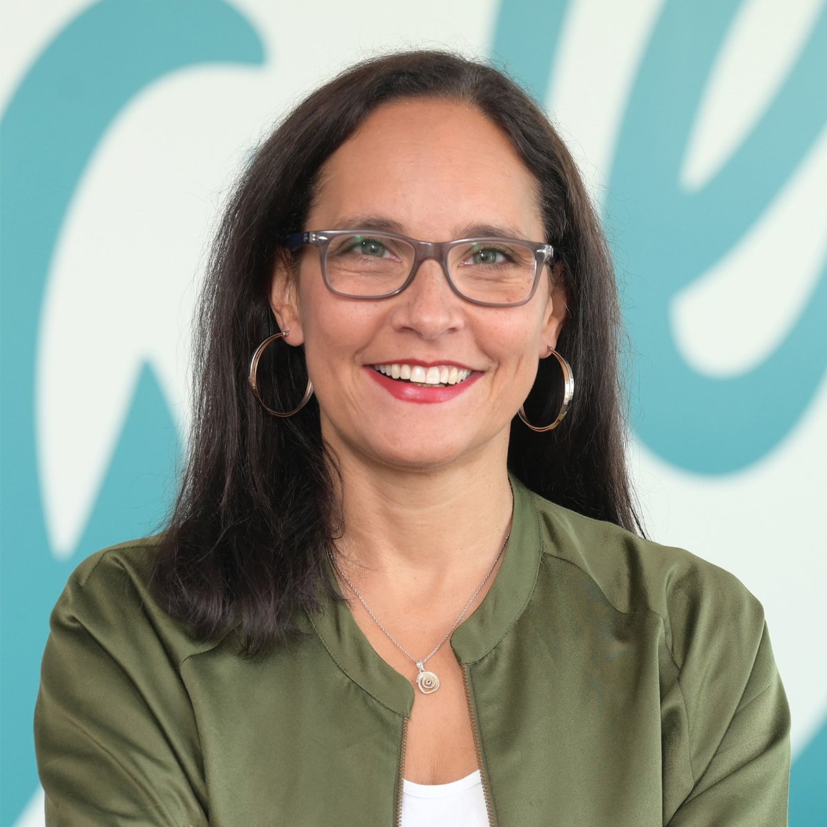 Andrea Spielmann: Eine Frau mit dunklem, schulterlangem Haar, Brille und roten Lippen lächelt freundlich in die Kamera. Sie trägt große Creolen-Ohrringe, eine Halskette und einen Ring. Ihr Outfit besteht aus einer weißen Bluse und einer olivgrünen Bomberjacke. Ihre Arme sind vor dem Körper verschränkt. Im Hintergrund ist eine helle Wand mit einem großflächigen türkisfarbenen Schriftzug unscharf zu erkennen. Die Szene wirkt freundlich und professionell.