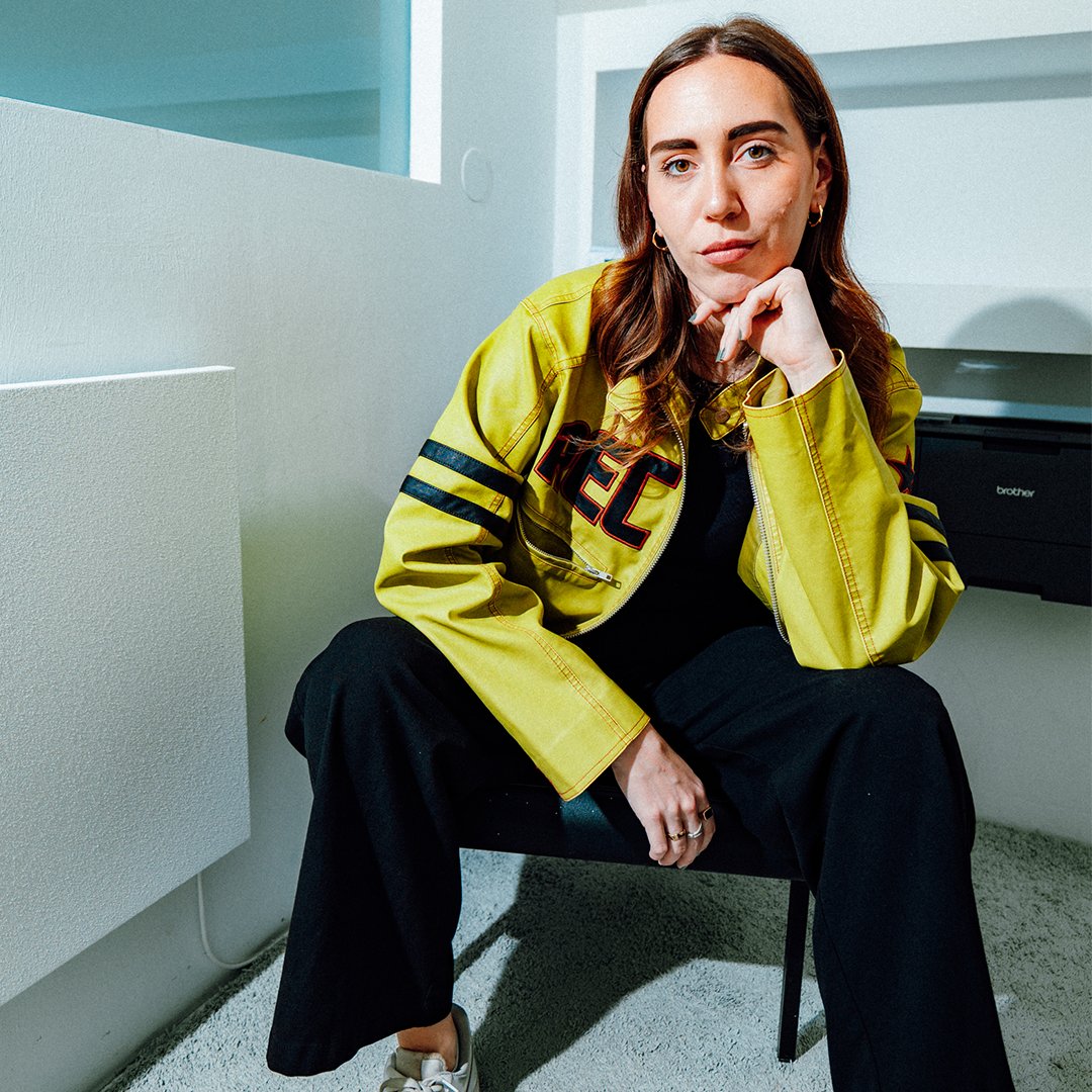 Anna Sramek sitting on a chair in a bright office setting, wearing a yellow jacket and looking directly at the camera.