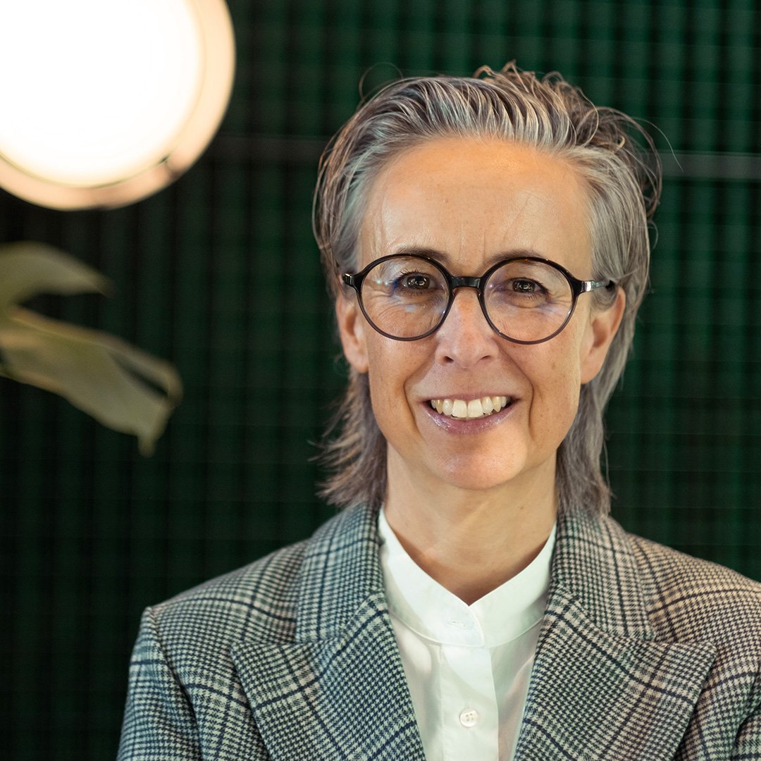 Porträt einer Frau mit kurzen grauen Haaren und runder Brille, die in die Kamera lächelt. Sie trägt ein kariertes Sakko und ein weißes Hemd, im Hintergrund sind Pflanzen und warmes Licht zu sehen.