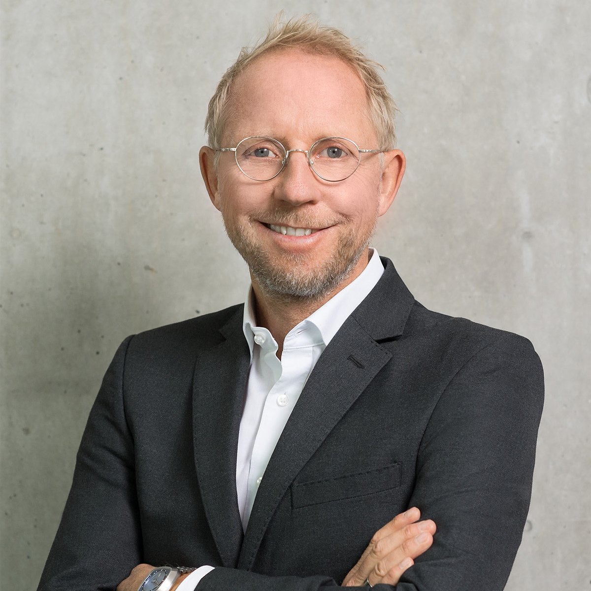 Bernd Rützler: Ein Mann mittleren Alters mit kurzem, blondem Haar und rundem Brillengestell steht vor einer grauen Betonwand. Er trägt ein dunkles Sakko über einem weißen Hemd, hat die Arme verschränkt und lächelt freundlich in die Kamera. Der Hintergrund ist schlicht und modern.