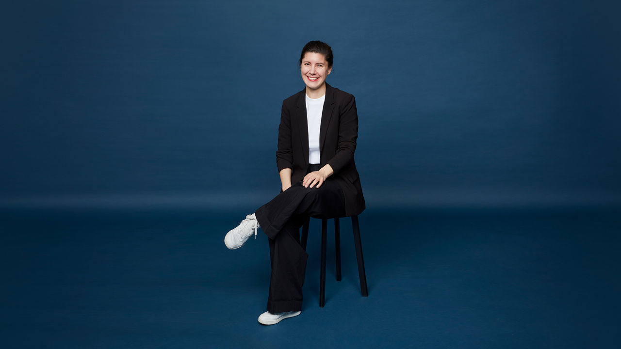 A woman with dark hair tied back sits smiling on a stool in front of a blue background, wearing a white T-shirt, a black blazer, wide black pants, and white sneakers, with her legs crossed casually.