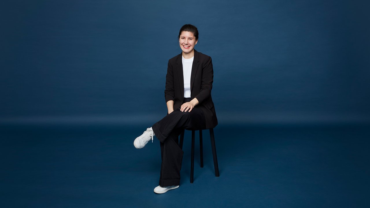 Eine Frau mit dunkel zusammengebundenem Haar sitzt lächelnd auf einem Hocker vor einem blauen Hintergrund, trägt ein weißes T-Shirt, einen schwarzen Blazer, schwarze weite Hose und weiße Sneakers, und hat die Beine locker übereinandergeschlagen.