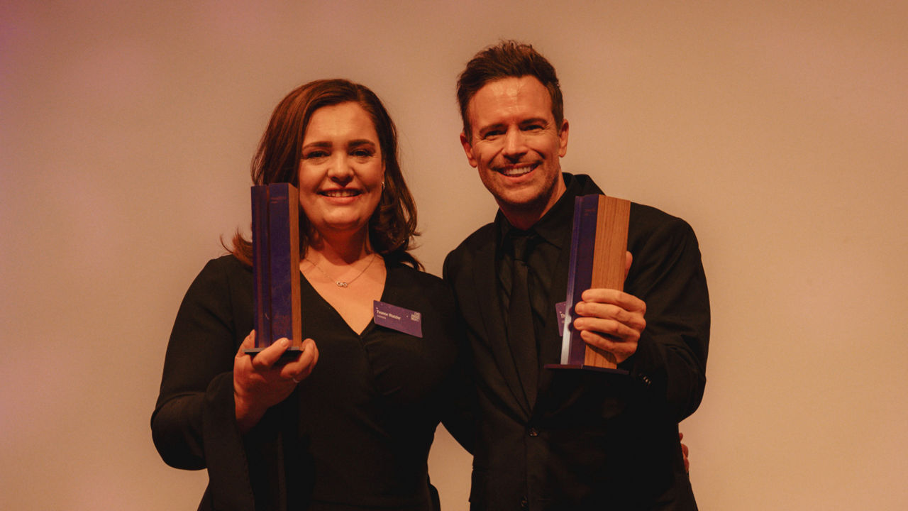 Yvonne Wutzler und Thorsten Mühl stehen lächelnd nebeneinander auf einer Bühne vor hellem Hintergrund und halten jeweils eine Trophäe in der Hand. Beide tragen schwarze Kleidung und Namensschilder. Die Szene zeigt die Auszeichnung zum „CMO of the Year 2025“. Das Bild vermittelt einen feierlichen Moment der Anerkennung für herausragende Marketingleistungen.