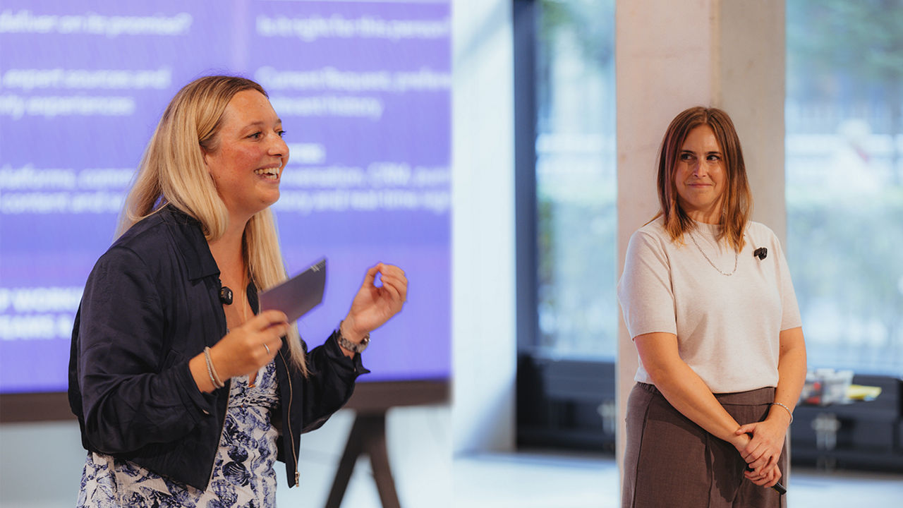 Zwei Personen (Carina Müller & Sophie von dem Bussche) stehen während einer Präsentation vor einer großen Bildschirmfläche. Die Person links hält eine Karte in der Hand und gestikuliert, während die Person rechts danebensteht. Beide tragen kleine Mikrofone. Im Hintergrund ist eine großformatige Präsentationsfolie in Blau- und Violetttönen mit mehreren Textabschnitten zu sehen.