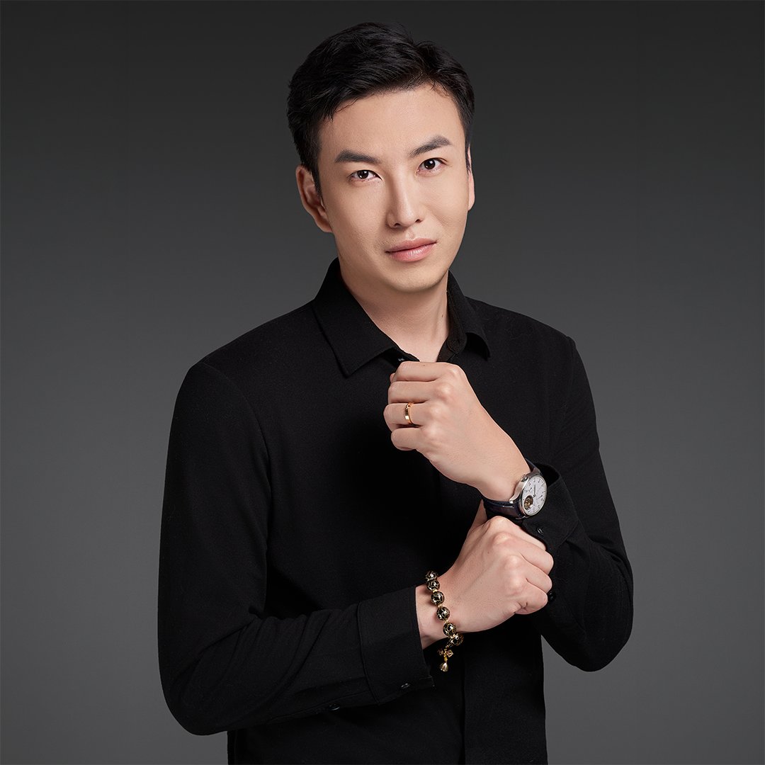 Professional studio portrait of a person wearing a black shirt in front of a dark grey background. The person is facing the camera with one hand near the collar. A wristwatch and bracelet are visible on the wrists. The image is presented in a formal portrait style.