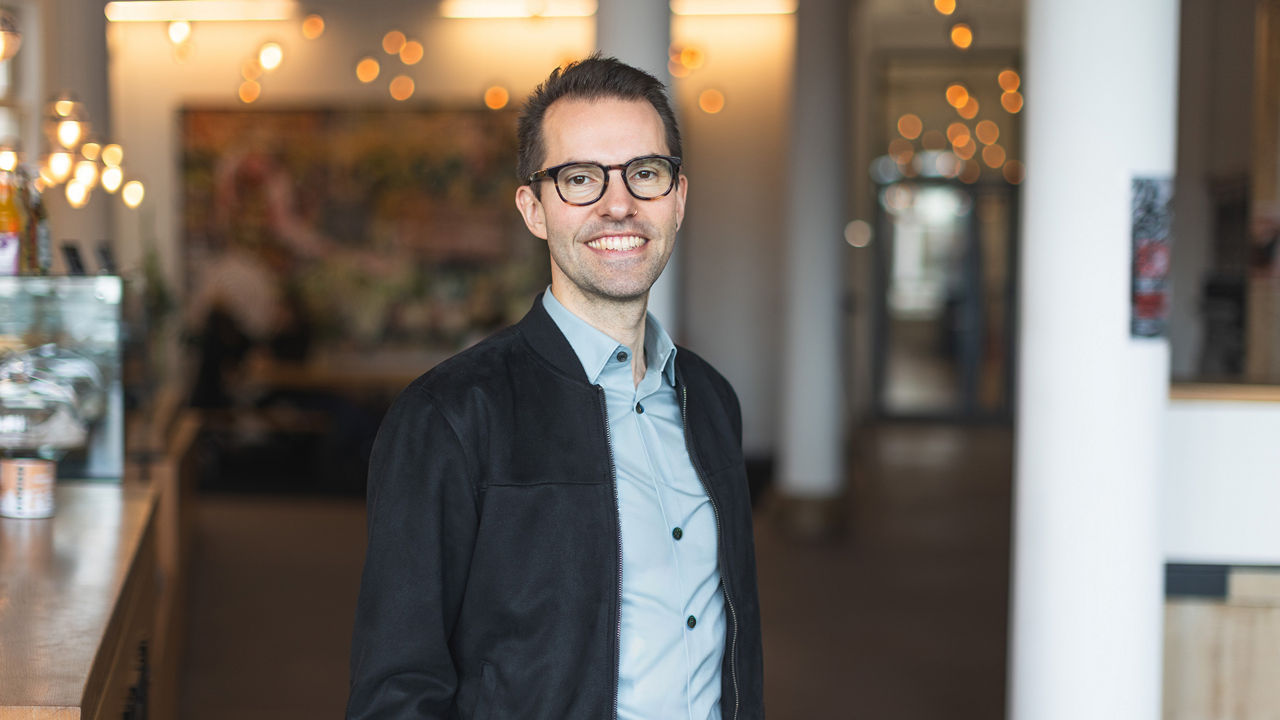 Im Vordergrund des Bildes steht ein lächelnder Mann (Chris Schneider) mit dunklem, kurzem Haar und Brille. Er trägt ein hellblaues Hemd und eine schwarze Jacke. Die Person befindet sich in einem modernen, offen gestalteten Innenraum mit warmem Licht und unscharfem Hintergrund, in dem sich Lichtreflexionen und Einrichtungsdetails wie ein Tresen und Lichterketten erkennen lassen. Die freundliche Ausstrahlung und die natürliche Pose der Person vermitteln Offenheit und Zugänglichkeit.