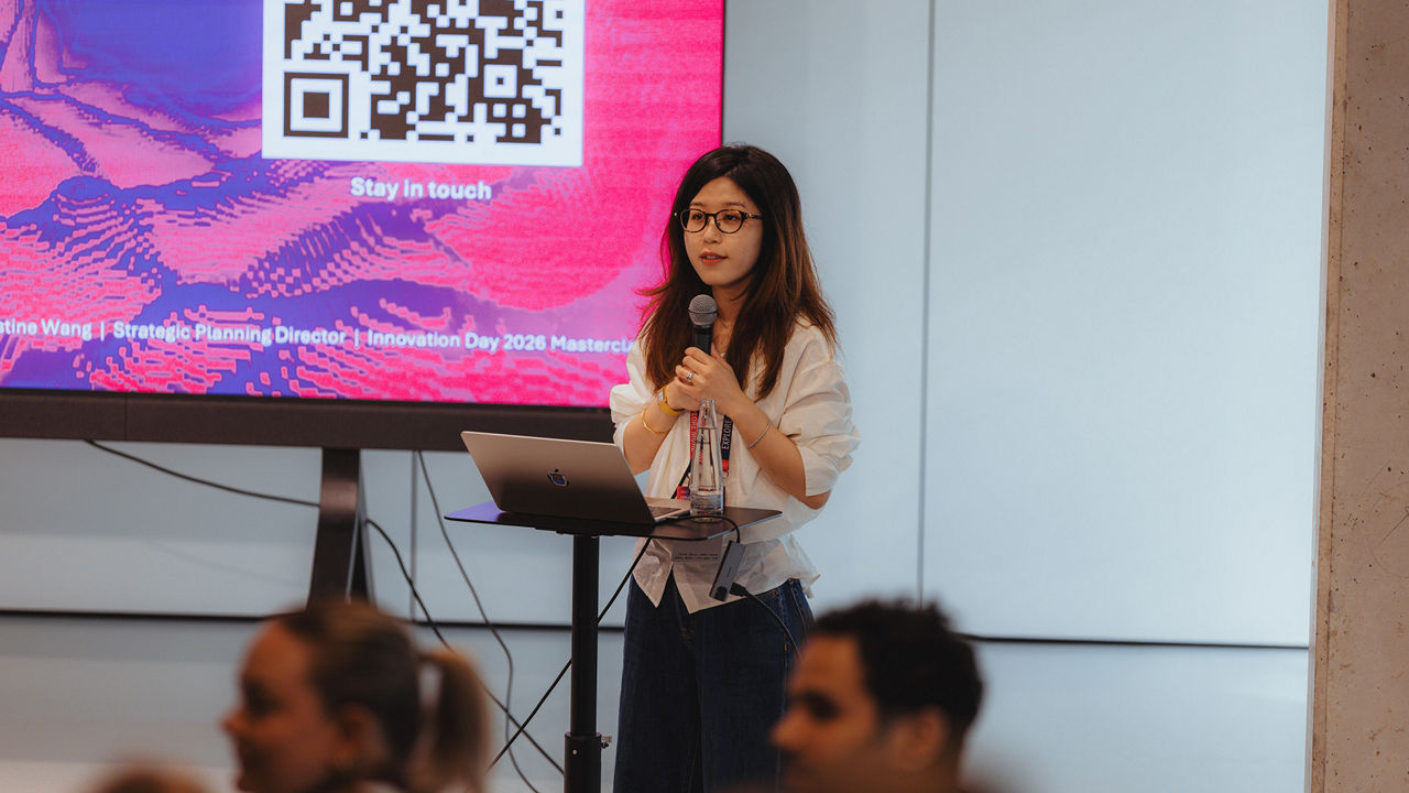 Christine Wang is standing next to a small table with a laptop and holding a microphone during a presentation. A large screen displaying a slide with a QR code and graphic elements is visible in the background. Audience members can be seen in the foreground. The image shows a conference or workshop presentation in a professional event setting.