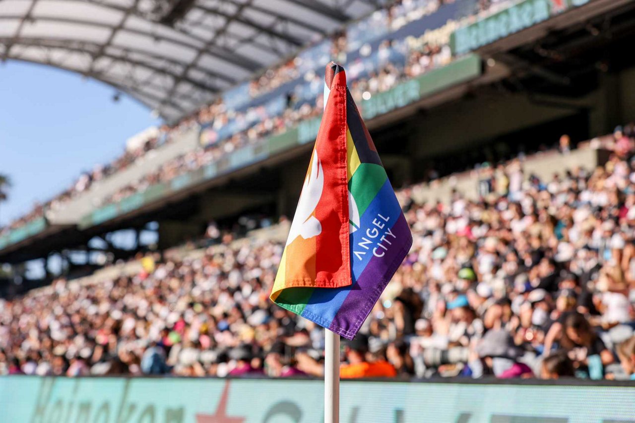 Eine Regenbogen-Eckfahne mit der Aufschrift „Angel City“ steht am Spielfeldrand eines Fußballstadions. Im Hintergrund sind die Tribünen mit zahlreichen Zuschauerinnen und Zuschauern zu sehen.