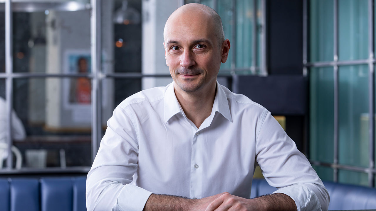 Portrait of a man wearing a white shirt, seated at a table in a modern office or lounge setting, looking directly at the camera.