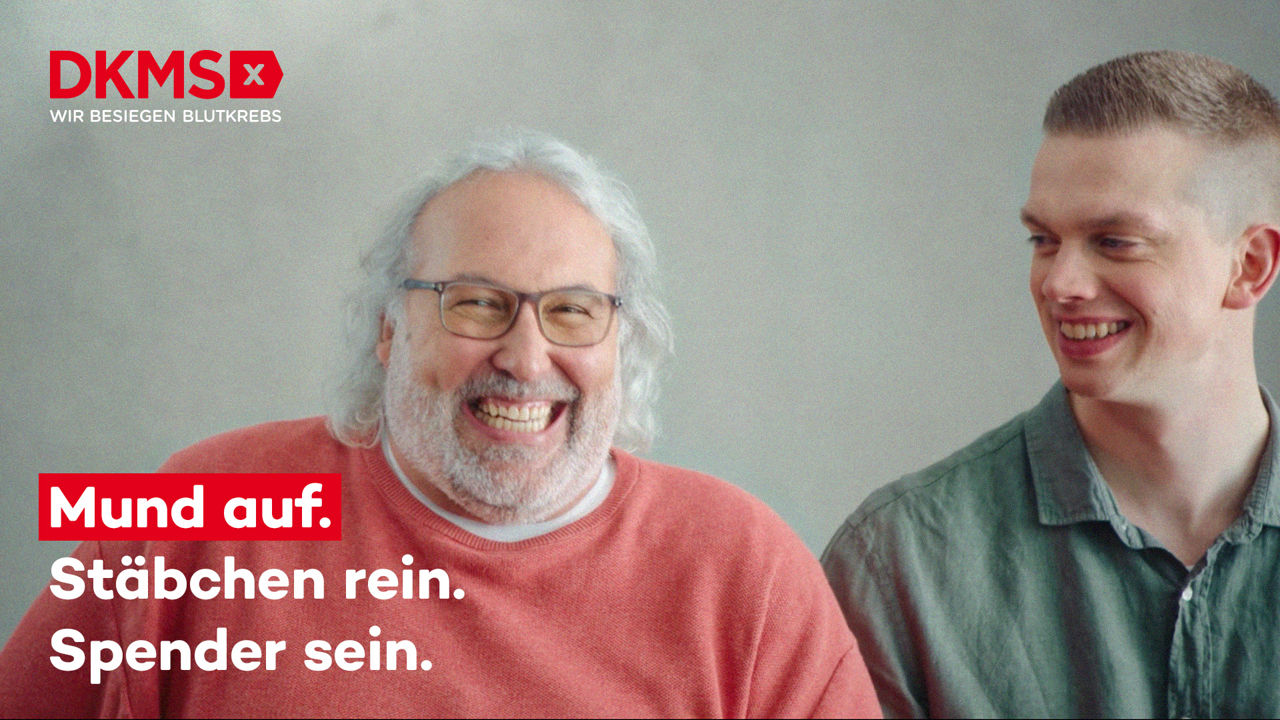 Zwei lachende Männer sitzen nebeneinander vor einem neutralen Hintergrund; links oben im Bild befindet sich das Logo der DKMS mit dem Slogan ‚WIR BESIEGEN BLUTKREBS‘ und darunter der Aufruf: ‚Mund auf. Stäbchen rein. Spender sein.‘ in weißer und roter Schrift.“