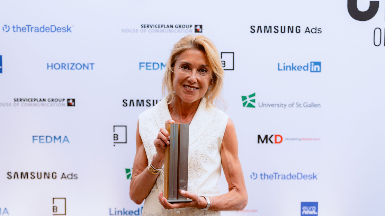 A person stands in front of a sponsor backdrop holding an award trophy. The backdrop features logos including Samsung Ads, LinkedIn, Serviceplan Group, The Trade Desk, FEDMA, Horizont, and the University of St. Gallen. The person is wearing a light-colored sleeveless top and looking at the camera.