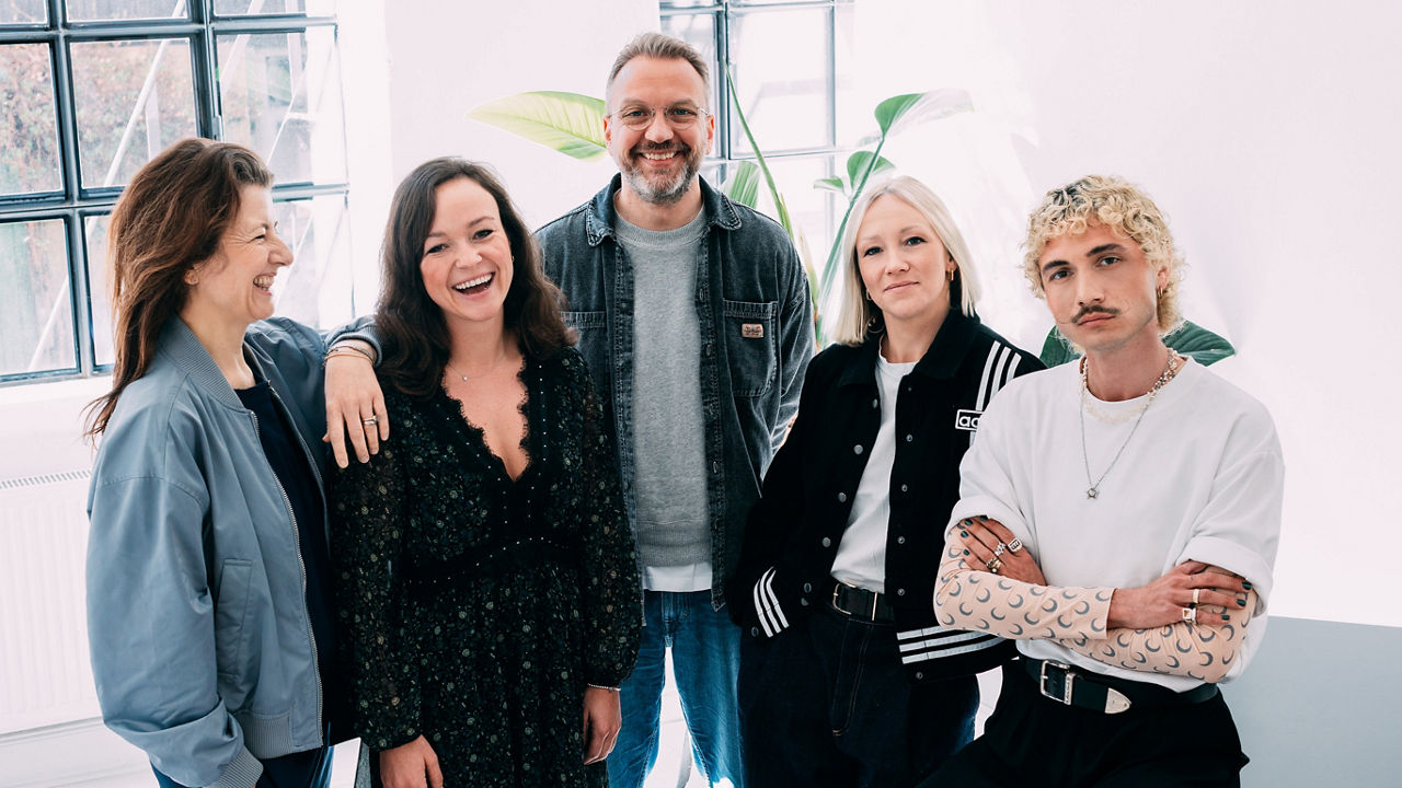 Fünf Personen in einem hellen Raum mit großen Fenstern und Pflanzen, stilvoll gekleidet und entspannt – einige lachend, andere mit cooler Pose. Zusehen sind auf dem Bild von links: Stefanie Kuhnhen (Serviceplan), Lill Brüggemann (Rügenwalder Mühle), Steffen Zeller (Rügenwalder Mühle), Madeleine Haug (Serviceplan Hamburg), Maurice Endres (Serviceplan Culture)