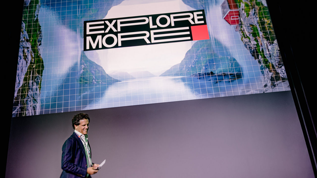 Person (Florian Haller) stands on a stage holding notes while a large projection screen above displays the words “EXPLORE MORE” against a graphic background featuring landscape imagery and grid elements. The image shows a presentation or conference setting.
