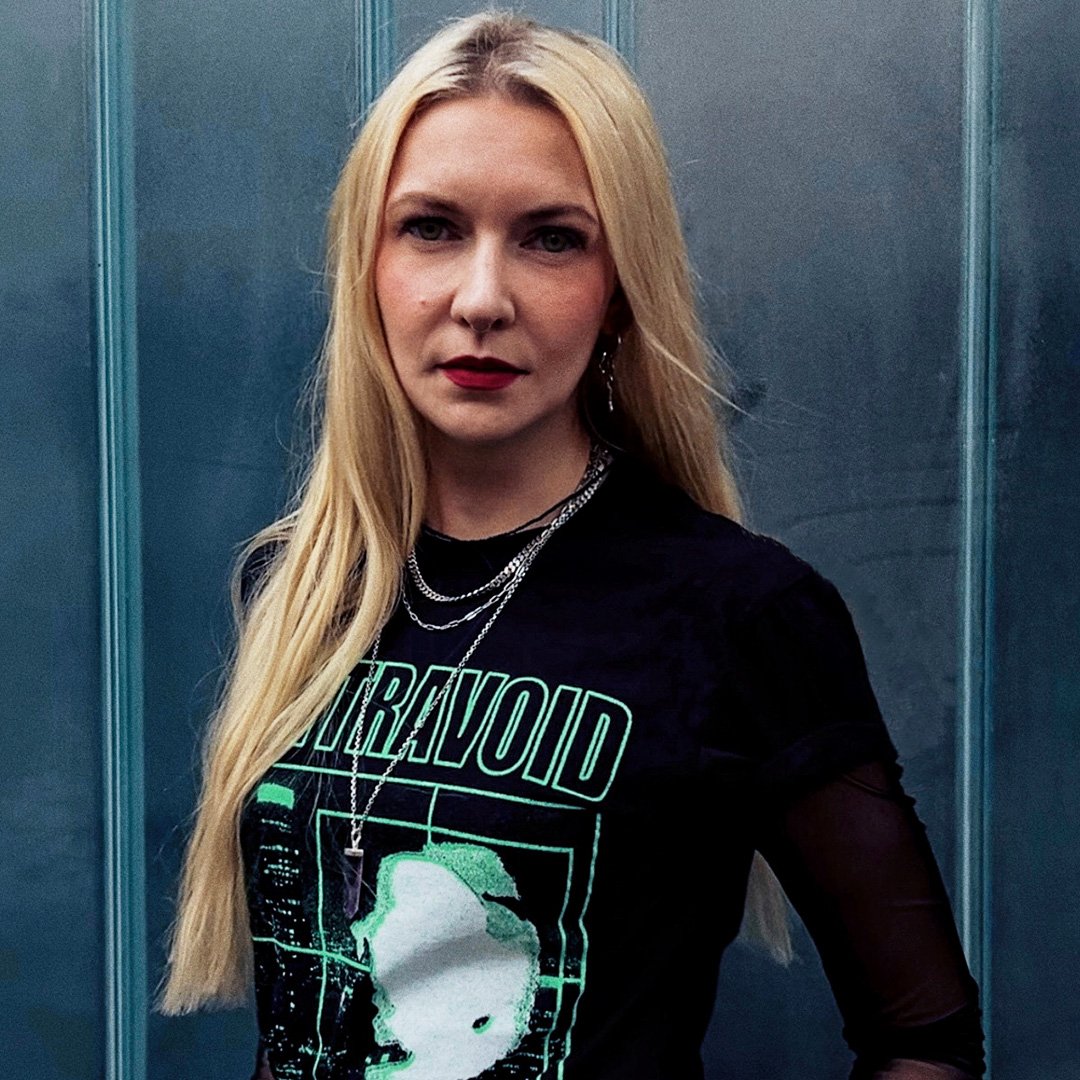 Porträt einer Frau mit langen blonden Haaren vor einer metallisch wirkenden Wand. Sie trägt ein schwarzes Shirt mit grafischem Print und blickt ernst direkt in die Kamera.