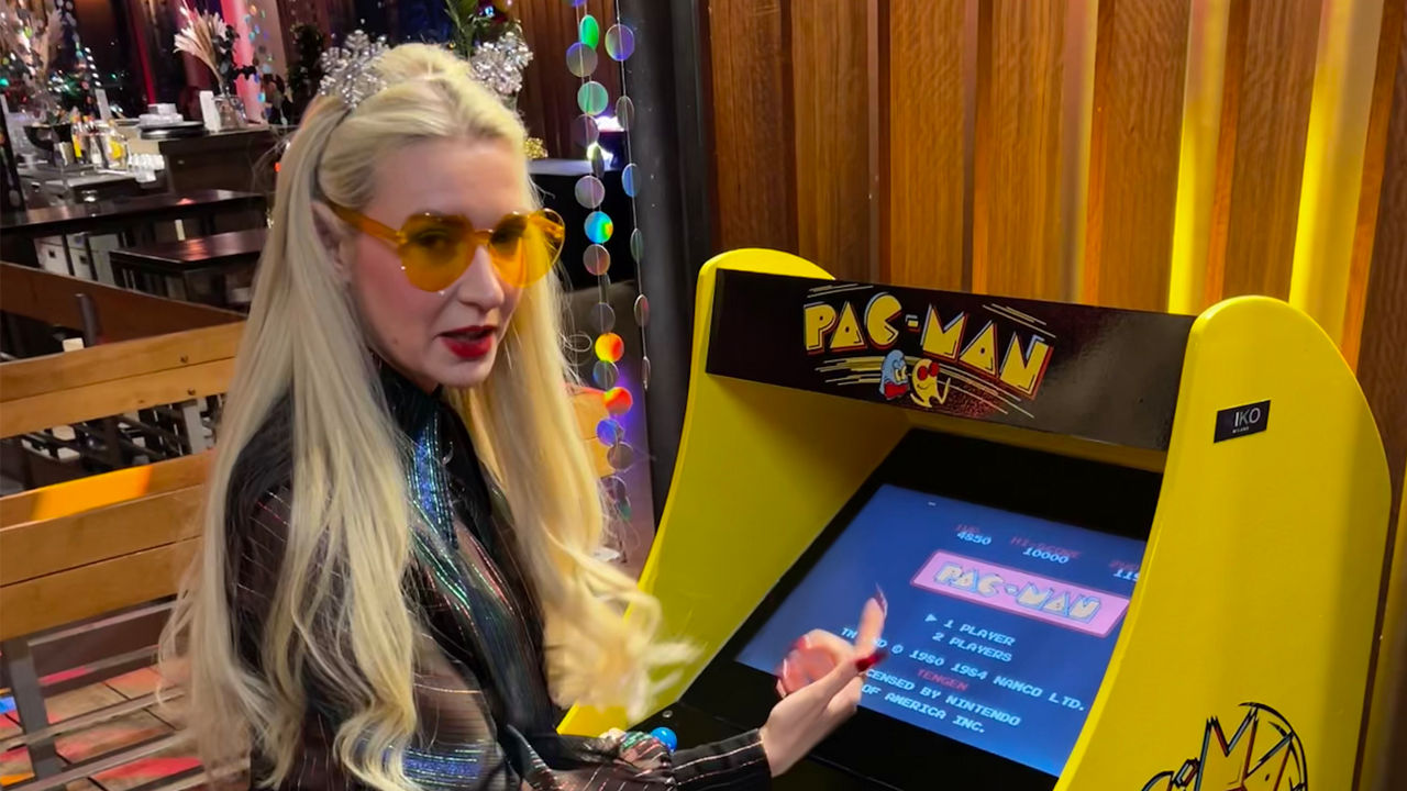 Person erklärt einen Pac-Man-Arcade-Automaten in einer dekorierten Innenumgebung.