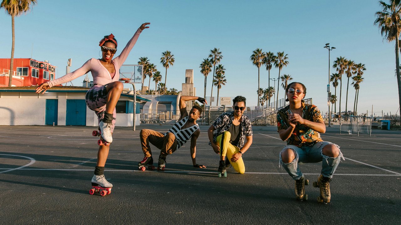 A group of five diverse young adults are roller skating and posing expressively on an outdoor court near a beach, surrounded by palm trees and bathed in warm sunlight, with clear skies and a vibrant atmosphere.