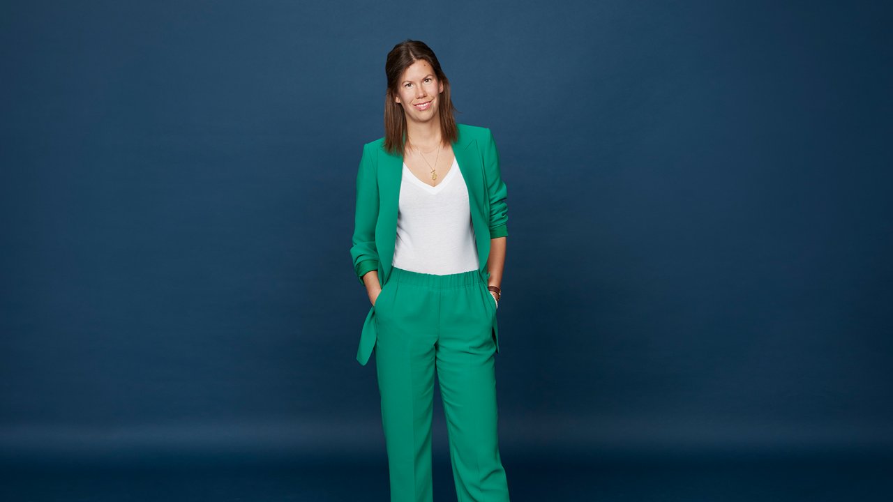 Professional portrait of a person wearing a teal pantsuit against a dark blue background.