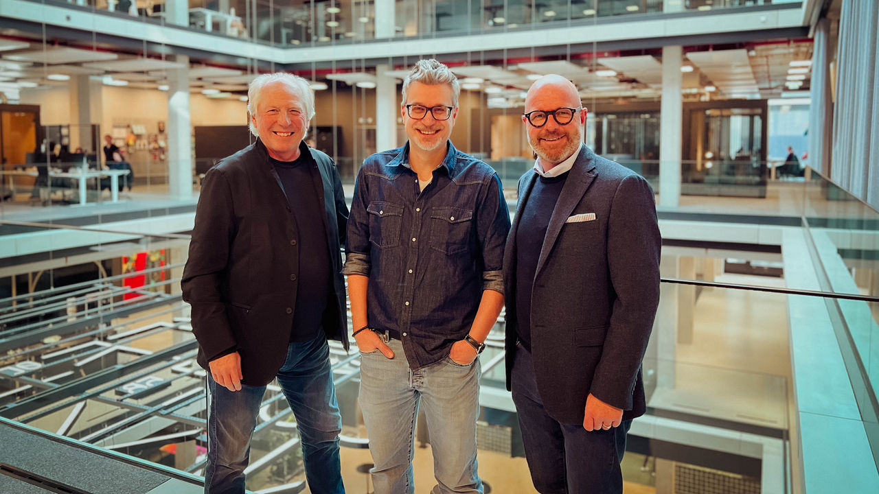 Drei erwachsene Personen stehen nebeneinander in einem modernen, lichtdurchfluteten Bürogebäude mit Glasfassaden und offenen Ebenen. Sie lächeln in die Kamera und tragen legere bis halbformelle Kleidung.