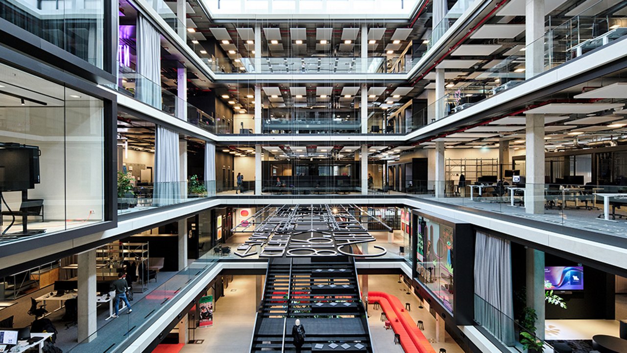 Bild des House of Communication in München: Modernes, offenes Bürogebäude mit mehreren Etagen, Glaswänden und einem zentralen Atrium mit Treppen und Sitzbereichen.