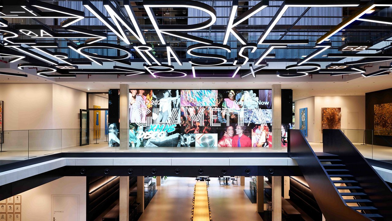 Modern multi-level atrium featuring a large LED video wall and a long central wooden table. A prominent illuminated ceiling installation.