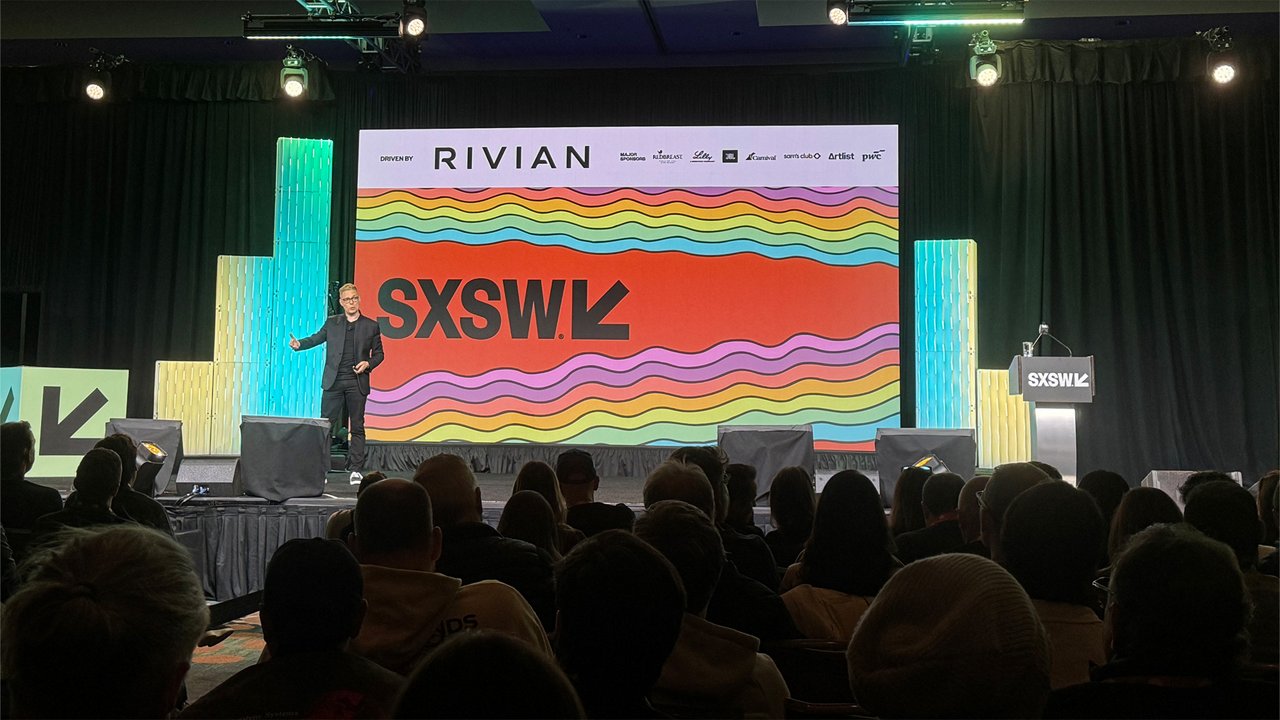 Person giving a presentation on a SXSW stage in front of a large screen with the SXSW logo and colorful background; audience seated in the foreground.