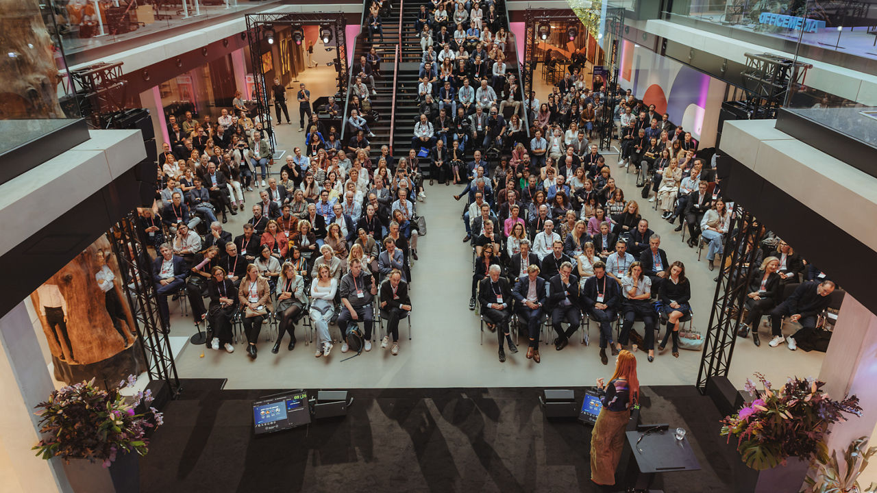 A large audience gathered in a multi-level event venue, facing a stage where a speaker addresses the crowd.