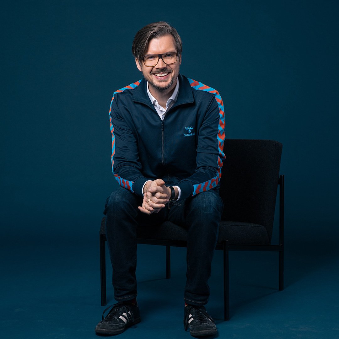 Mann mit Brille sitzt auf einem Stuhl vor blauem Hintergrund und lächelt in die Kamera. Er trägt eine sportliche Jacke und hat die Hände locker ineinander verschränkt.