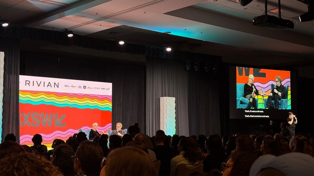 Two speakers sit on stage at SXSW in a discussion in front of an audience, with their conversation shown on a large screen.