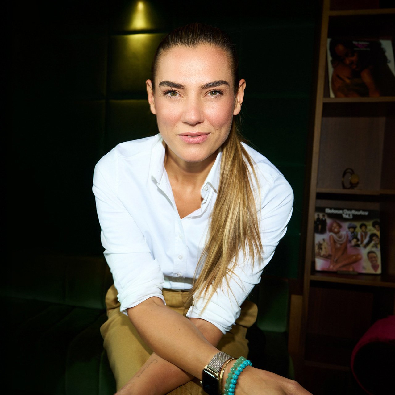 Kathrin Jesse, a woman with fair skin and long straight hair tied to the side, is seated in a warmly lit room. She is wearing a white shirt, beige trousers, and bracelets on her wrist. A shelf with vinyl records is visible in the background.