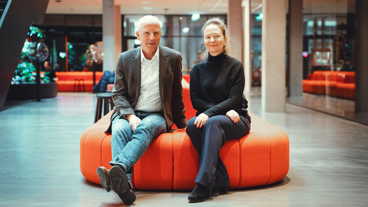 Zwei Personen sitzen nebeneinander auf einem runden, orangefarbenen Polstermöbel in einer modernen, offen gestalteten Lobby mit Glaswänden und warmem Licht.