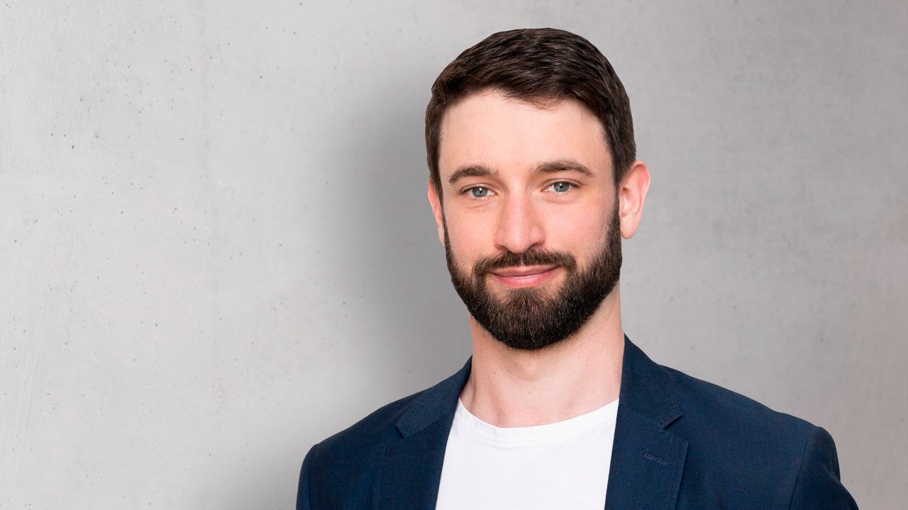 Kevin Heil, Unit Director at hmmh AG: The photo shows a smiling man with short brunette hair in a navy suit who looks directly into the camera.