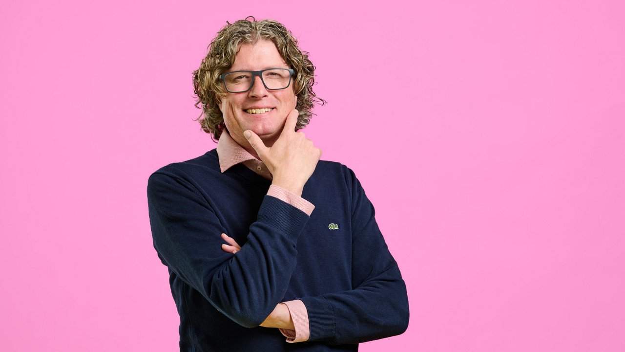 Pim vn Houten: Portrait of a man with medium-length curly hair and glasses against a pink background. He wears a dark blue sweater over a pink shirt. His arms are crossed, and one hand touches his chin while he smiles warmly at the camera.