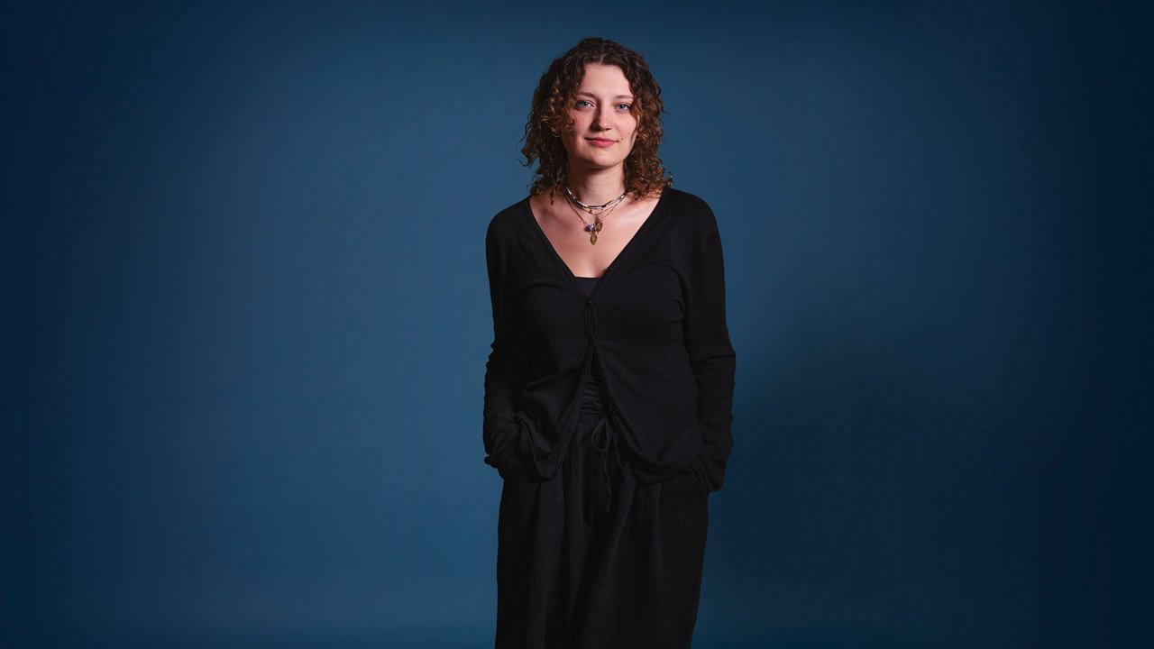 A woman with curly, shoulder-length brown hair stands in front of a blue background, wearing a black outfit with a cardigan and layered necklaces, her hands casually in her pockets, and a subtle smile on her face.
