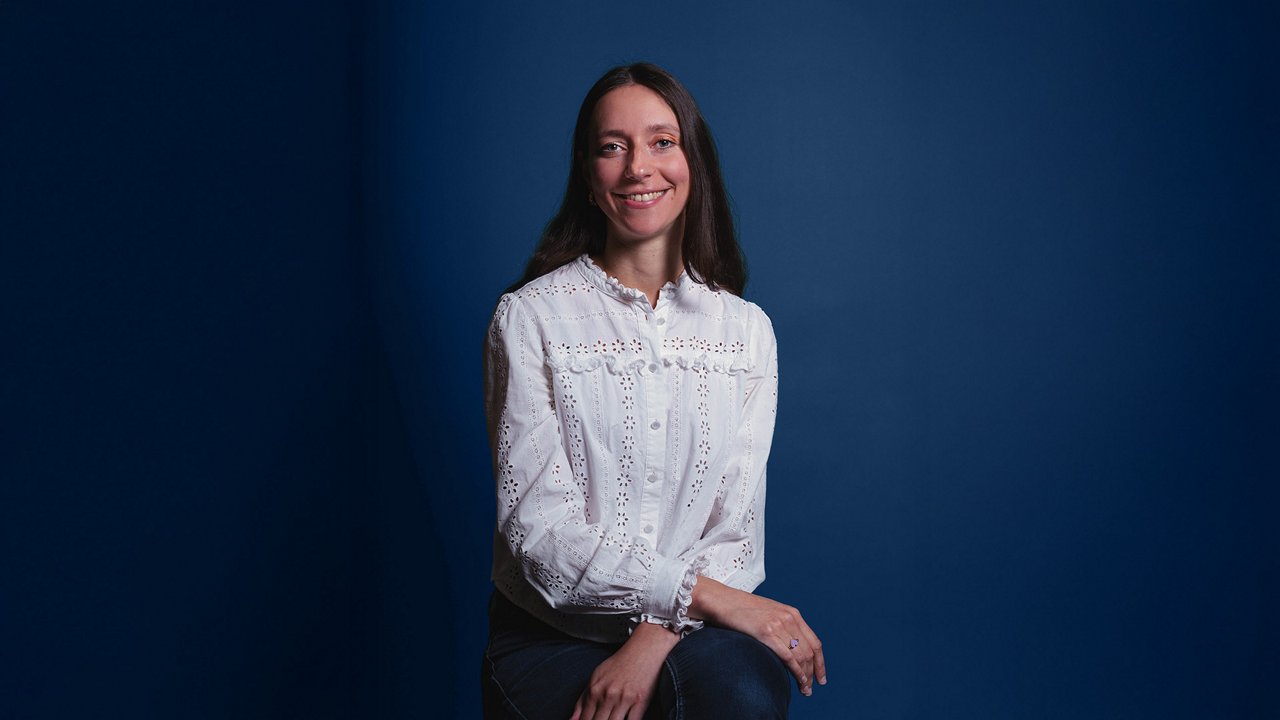 A woman with long dark hair sits in front of a plain dark blue background, wearing a white blouse with eyelet embroidery, smiling warmly at the camera with her hands resting loosely on her lap.