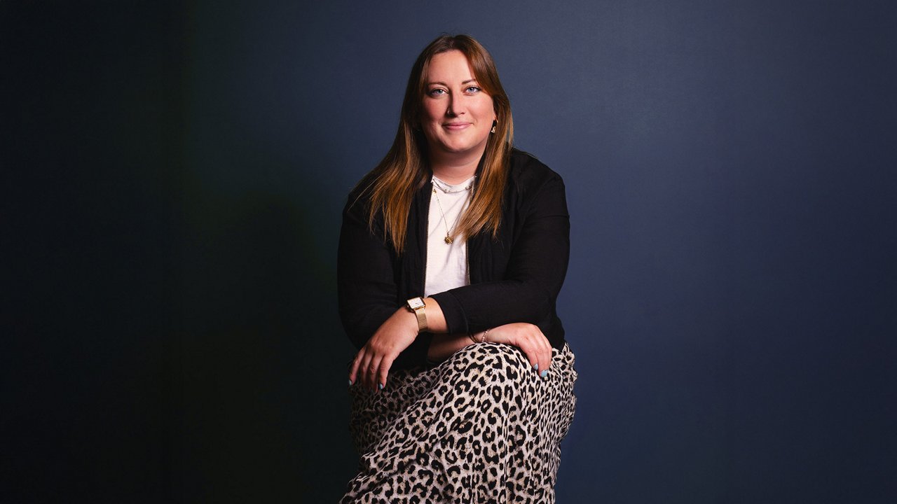 A woman with straight, shoulder-length light brown hair sits on a stool in front of a blue background, wearing a black outfit with layered necklaces and a watch, smiling with her hands resting loosely on her legs.