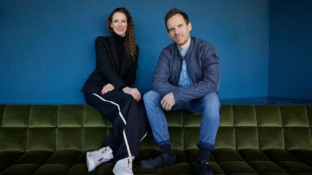 Eine Frau und ein Mann sitzen nebeneinander auf einem grünen Samtsofa vor einer blauen Wand. Die Frau trägt einen schwarzen Hosenanzug und hat lockiges, langes Haar. Der Mann trägt eine hellblaue Jeans, ein Jeanshemd und eine gesteppte Jacke. Beide lächeln in die Kamera.