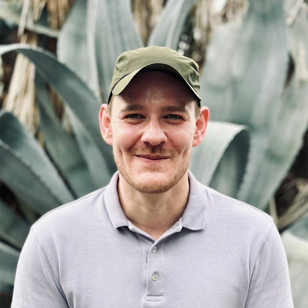 Lorenz Langgartner wearing a cap and polo shirt, smiling in front of large plants.