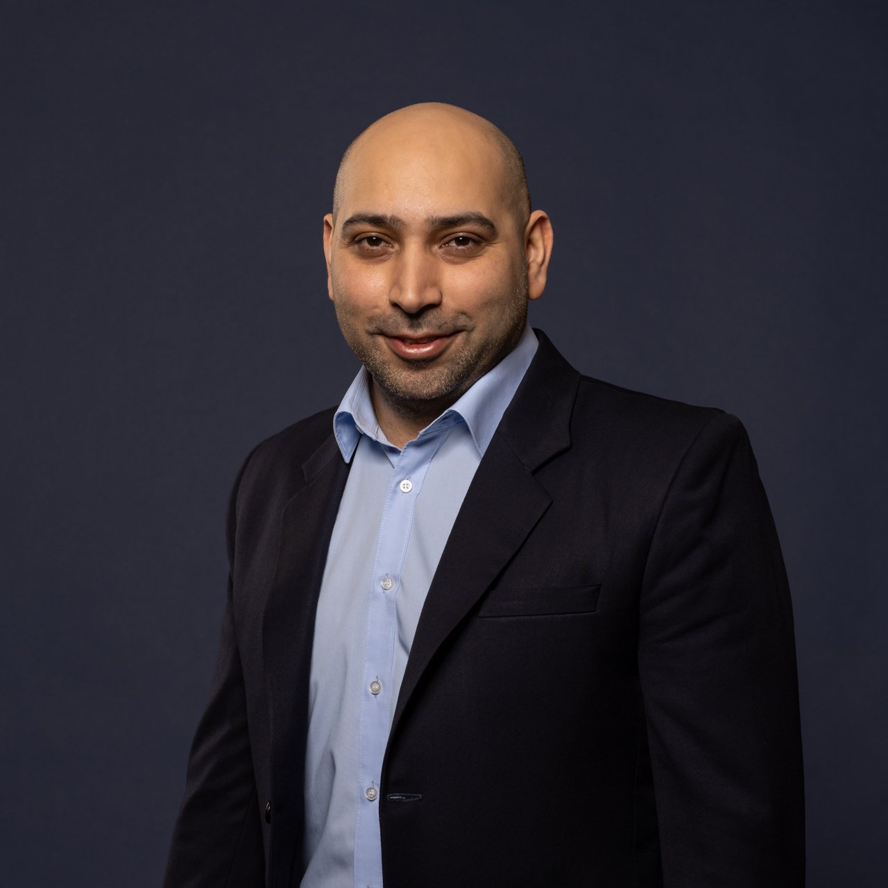 Lotfi Hadi, a man with a shaved head, medium skin tone and short beard, stands in front of a dark blue background. He is wearing a light blue shirt and a dark blue blazer, smiling gently at the camera.