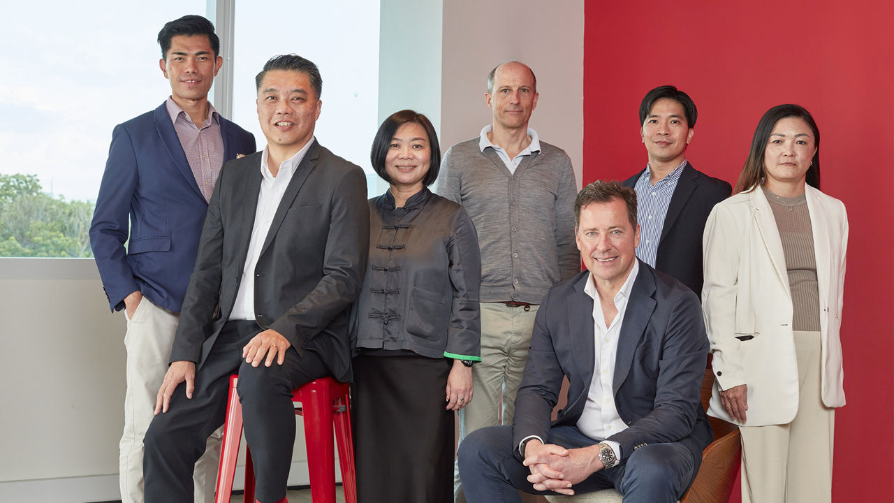 Group portrait of eight people (left to right: Jason Tan, Gary Tang, Jessica Toh, Julian Simons, Matthias Brüll, Joseph Lee, Ivy Rushbrook) in business attire in a modern office. Some are standing while two sit in front, with a window and a red wall visible in the background.
