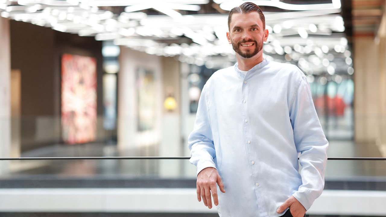 Uwe Roschmann: A man with a beard wearing a light blue shirt stands smiling by a railing in a modern building (House of Communication Munich) with bright ceiling lights in the background.