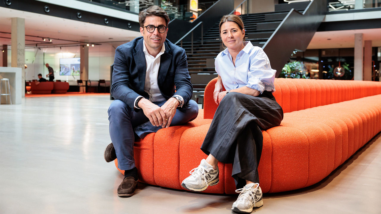 Christian Schmitz and Christin Herrnberger are seated on a modern, orange sofa in an open-plan office with exposed concrete ceilings and large windows.